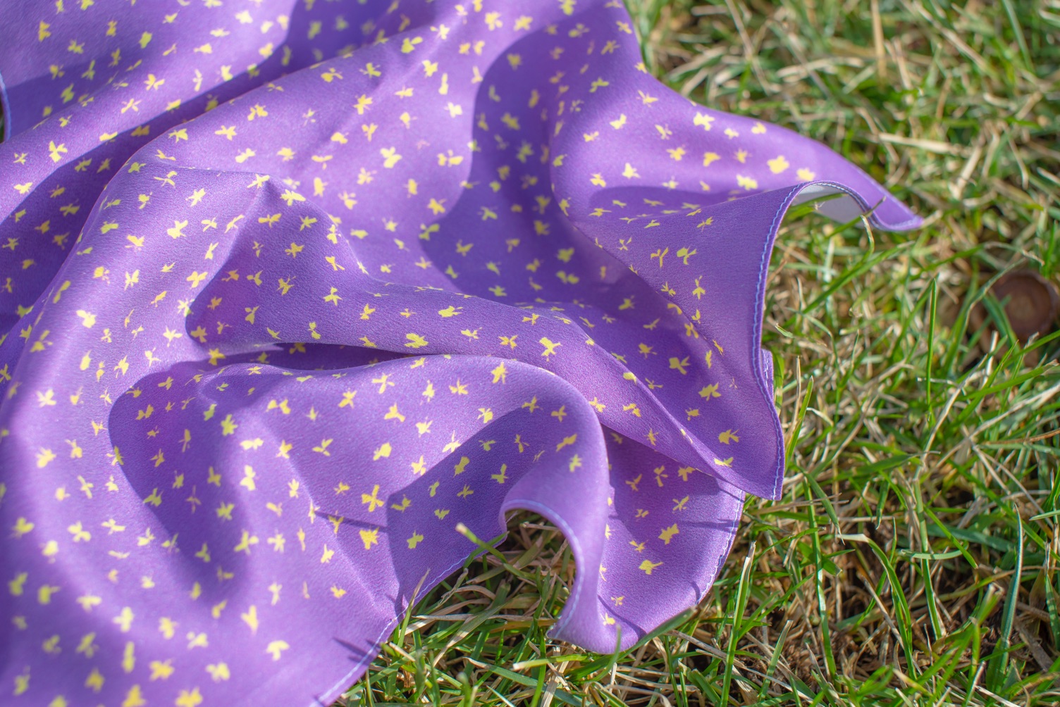 A silk handkerchief printed with a sprinkle of bright abstract flowers, generated from moments on my birthday, lays on the grass.